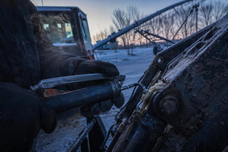 A worker greasing the pins of a tractor with a cordless grease gun on a frosty winter morning