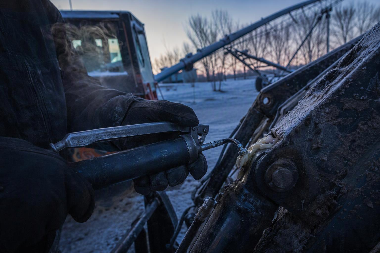 A worker greasing the pins of a tractor with a cordless grease gun on a frosty winter morning